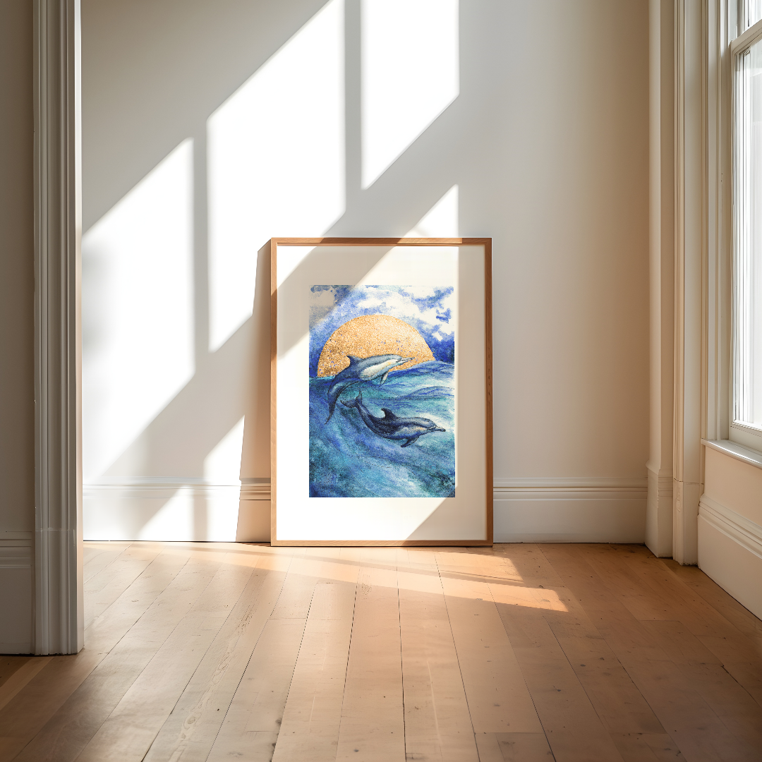 A framed art print depicting two dolphins in blue ocean waves with a golden sun, leaning against a wall on a light wood floor in a sunlit room.