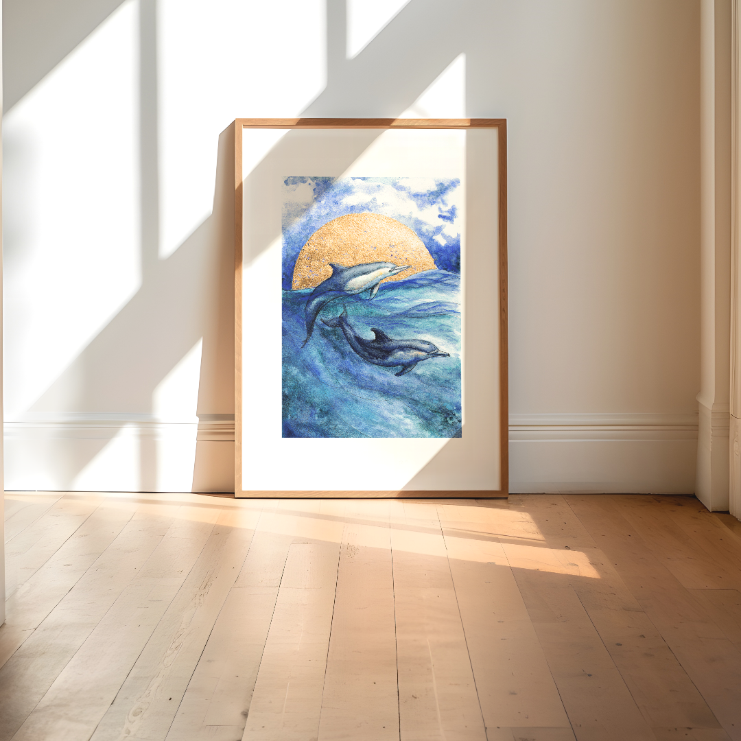A framed art print depicting two dolphins swimming in blue ocean waters under a golden sun, leaning against a white wall on a light wood floor with natural window shadows.